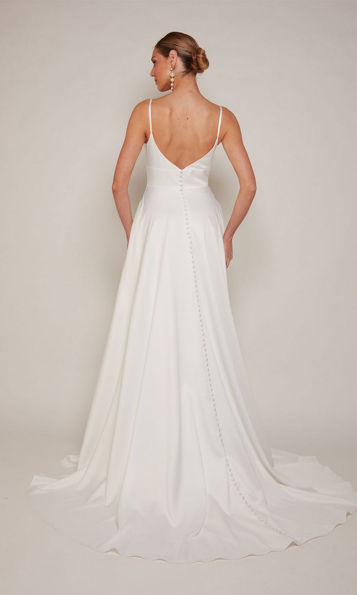 A woman in the Alyce 7095 long white dress, featuring a V-back and button detailing, stands facing away with her hair in a low bun.
